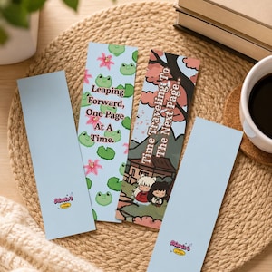 May include: Four rectangular bookmarks. Two are light blue with a small logo at the bottom. One features green pixelated frogs and pink flowers with the text Leaping Forward, One Page At A Time. One shows an anime scene with the text Time Traveling To The Next Page.