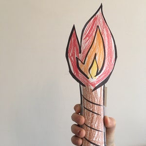 May include: A hand holds a paper craft of a torch. The torch is made of brown paper with a red, orange, and yellow flame drawn on top.