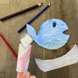 May include: A paper craft set for a children's story about a person being swallowed by a fish. The set includes a blue fish with a large open mouth, a small brown boat, and a person with red clothing.