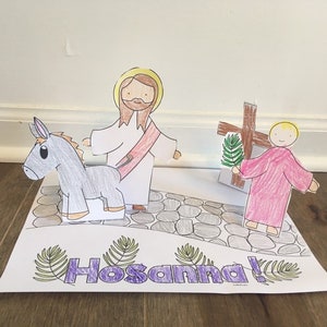 May include: A handmade paper craft depicting a religious scene. It features a figure riding a donkey, another figure, and a cross. The word "Hosanna!" is written in purple with green leaf accents. The figures are on a stone path.