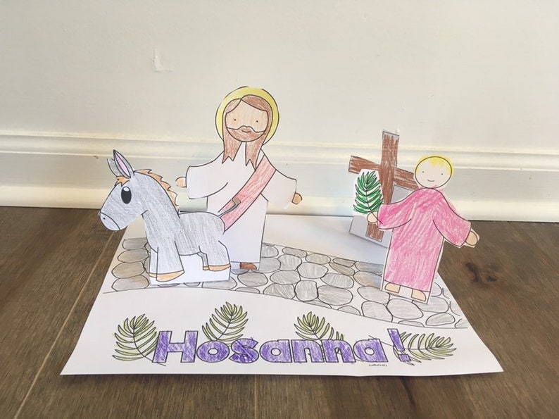 May include: A handmade paper craft depicting a religious scene. It features a figure riding a donkey, another figure, and a cross. The word "Hosanna!" is written in purple with green leaf accents. The figures are on a stone path.