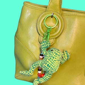 May include: A lime green leather handbag featuring a hanging accessory. The accessory consists of a bright green metal ring, a matching green plastic clasp, and a plush frog with a green and yellow spotted pattern, accented by a string of colorful beads.