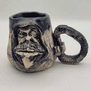 May include: A ceramic mug featuring a sculpted, hooded face with a mustache and goatee. The mug has a dark, textured finish with lighter, off-white highlights on the facial features. It includes a thick, rounded handle with a rough, pebbled texture.