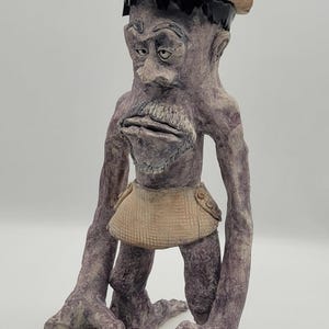 May include: A hand-sculpted clay figurine of a tall, purple-toned creature with long arms and sharp claws. The figure wears a tan cowboy hat, a black headband, and a textured tan loincloth. It has a prominent nose, a mustache, and a goatee.