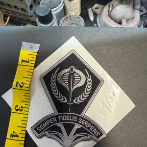 May include: A black and silver embroidered patch featuring a cobra and sword emblem with the text Semper Fidelis Serpens. A yellow measuring tape is placed alongside the patch, indicating a height of approximately 4.5 inches for the design.