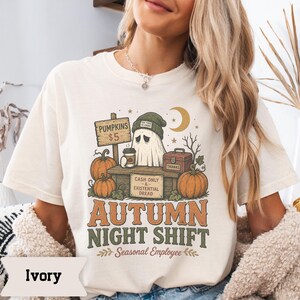 May include: An ivory t-shirt featuring a graphic of a ghost wearing a green beanie at a pumpkin stand. The design includes text reading PUMPKINS $5, CASH ONLY & EXISTENTIAL DREAD, THANKS, AUTUMN NIGHT SHIFT, and Seasonal Employee with pumpkins.