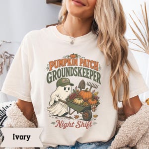 May include: An ivory t-shirt featuring a graphic of a ghost wearing a green cap, pushing a wheelbarrow filled with pumpkins and autumn leaves. The text reads PUMPKIN PATCH GROUNDSKEEPER and Night Shift, with a shovel and rake in the background.