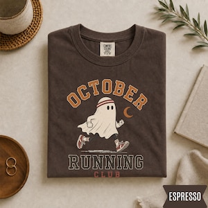 May include: A dark brown t-shirt featuring a graphic of a white ghost wearing a headband and sneakers, running. The text reads OCTOBER RUNNING CLUB in orange and white block letters. A small orange crescent moon is visible next to the ghost.