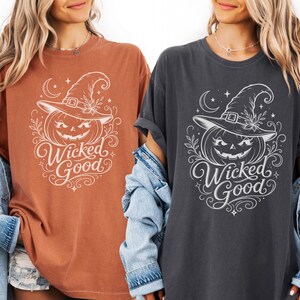 May include: Two short-sleeved t-shirts, one in rust orange and one in charcoal gray, each featuring a white graphic print of a jack-o'-lantern wearing a witch hat, surrounded by stars and swirls, with the text Wicked Good written below.