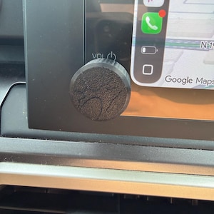 May include: A circular black volume knob cover with a textured, tree-like pattern on its face. The knob is attached to the volume control dial on a vehicle's infotainment screen, which displays a Google Maps interface with navigation icons.