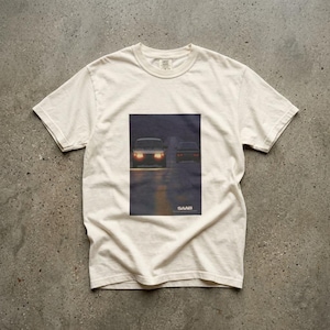 May include: An off-white short-sleeve t-shirt featuring a rectangular graphic print on the chest. The print depicts two cars driving on a dark road at night with headlights illuminated. The word SAAB is printed in white at the bottom of the graphic.