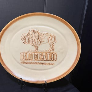 May include: An oval ceramic platter with a cream-colored base and a brown rim. The center features an embossed illustration of a buffalo standing above the text "BUFFALO CONSTRUCTION, INC" in raised, brown-toned lettering.