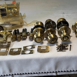 May include: A collection of brass-toned door hardware including several round knobs, a lever handle, latch mechanisms, rectangular strike plates, and a small pile of metal screws. Some components are still sealed in clear plastic packaging.