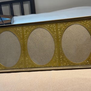 May include: A rectangular gold-toned metal picture frame featuring three oval-shaped openings. The frame has a textured, embossed floral pattern surrounding the oval cutouts and a thin, smooth metallic border along the outer edges.