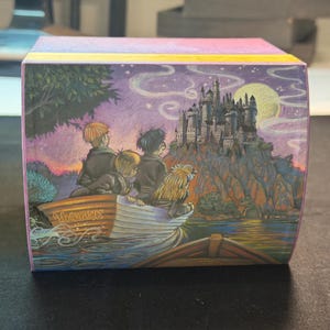 May include: A square box featuring a colorful illustration of three figures in a small boat on water, looking toward a large castle on a cliff under a full moon. The side of the boat is inscribed with the word Hogwarts in a gold-colored script.
