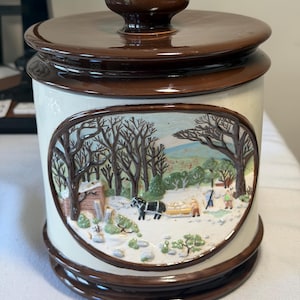 May include: A cylindrical ceramic canister with a glossy brown lid and base. The cream-colored body features a raised, oval-shaped relief depicting a winter scene with bare trees, a horse-drawn sled, and figures in a snowy landscape.