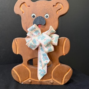 May include: A brown wooden bear cutout with googly eyes, a black painted nose, and a small red mouth. The bear features a cutout in the center of its torso and is decorated with a cream-colored ribbon featuring a repeating blue bear pattern.
