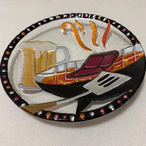 May include: A round glass plate featuring a painted design of a beer mug, a grilled steak, and a spatula. The rim is decorated with a black border containing alternating orange and silver dots. Steam lines rise from the steak, and the background is clear.