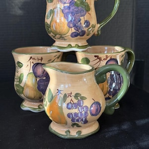 May include: A set of three ceramic mugs and one creamer pitcher, all featuring a light tan base with hand-painted illustrations of yellow pears, purple grapes, and green leaves. Each piece has a scalloped base and a solid green handle.