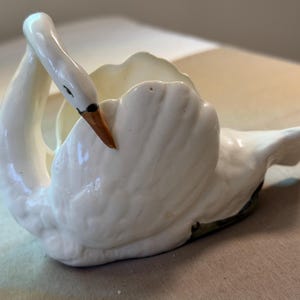 May include: A white ceramic swan figurine with a glossy finish, featuring a long, curved neck and a beak painted in a light brown hue. The swan is depicted in a resting position with its wings folded, creating a hollow center for storage.