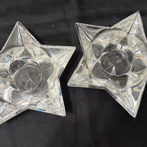 May include: Two clear glass candle holders shaped like five-pointed stars. Each holder features a circular center depression designed to hold a candle, with faceted edges that catch the light and create a geometric, star-shaped silhouette.