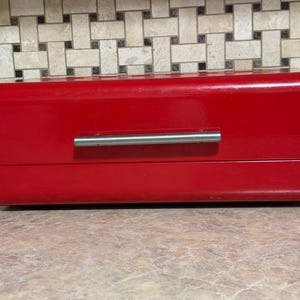 May include: A rectangular, glossy red bread box featuring a horizontal, brushed silver metal handle centered on the front panel. The container has a smooth, solid-colored finish and a clean, minimalist design with rounded edges on the top and bottom.