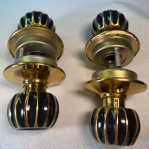 May include: Two door knobs featuring round, fluted black centers with gold-toned metal accents. Each knob has a circular gold-toned mounting plate and a central spindle mechanism extending from the back, showcasing a metallic finish throughout.