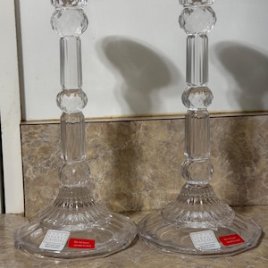 May include: A pair of clear crystal candlesticks featuring a faceted spherical design on the stem, a flared base, and a wide top rim. Each base displays a white label reading Fifth Avenue Crystal 24% Lead Crystal Made in Austria and a red label.