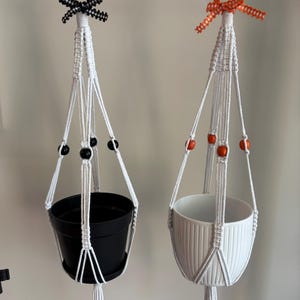 May include: Two white macrame plant hangers with knotted cord designs. One hanger features black beads and a black and white gingham ribbon, holding a black pot. The other features orange beads and an orange gingham ribbon, holding a white ribbed pot.