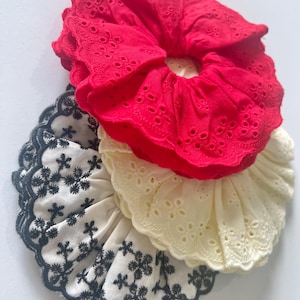 May include: Three fabric hair scrunchies featuring ruffled edges with eyelet embroidery. One scrunchie is bright red, one is off-white, and the third is white with a black floral-style embroidered pattern along the scalloped edge.