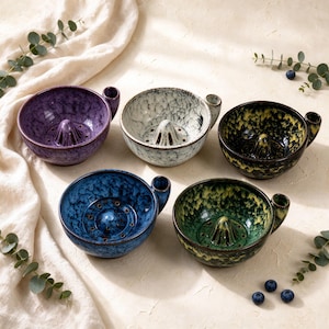 May include: Five ceramic citrus juicers in purple, white, black and yellow, blue, and green and yellow. Each bowl features a central reamer, a small side handle, and a speckled glaze finish. The juicers are arranged in a group on a light surface.