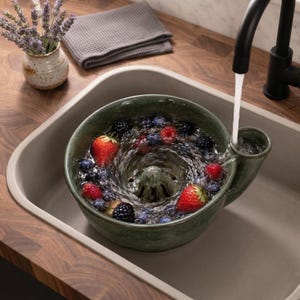 May include: A dark green ceramic berry colander bowl with a central drainage cone and a side spout. The bowl is filled with water and fresh strawberries, blueberries, and blackberries, while a stream of water flows from a tap into the spout.