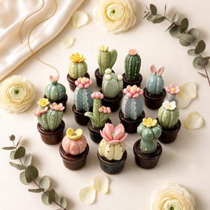 Pode incluir: Uma coleção de pequenas figuras de cactos em cerâmica em vasos castanhos escuros. Cada cato apresenta formas únicas, tons de verde, azul ou rosa, e é rematado com delicadas flores esculpidas ou pequenos detalhes como orelhas de coelho.