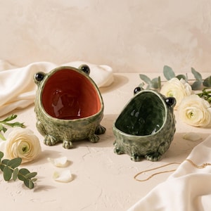 May include: Two ceramic frog-shaped bowls with textured green outer surfaces and black eyes. One bowl features a smooth, glossy orange interior, while the other bowl has a matching glossy green interior. Both bowls have small feet and wide openings.