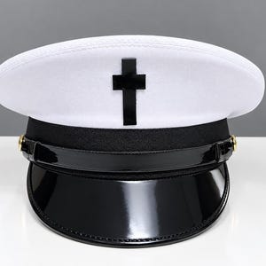 May include: A white peaked cap featuring a black cross emblem centered on the front crown. The hat includes a black band, a glossy black patent leather visor, and gold-toned buttons on each side securing a thin black chin strap across the front.