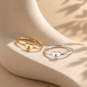 Può includere: Due sottili anelli in metallo, uno color oro e uno color argento, ciascuno caratterizzato da un piccolo ciondolo a forma di cuore. L'anello dorato è inciso con la lettera S, mentre l'anello argentato presenta la lettera K sui cuori.