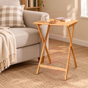 May include: A light brown bamboo folding snack table with a rectangular top. On the table sits a white ceramic mug with a floral pattern, a small white plate containing several round cookies, and a closed rectangular book with a light cover.