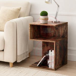 May include: A C-shaped side table with a dark brown wood grain finish. The table features an open storage compartment for books and a flat top surface. A small potted green plant and a silver metal desk lamp sit on the top of the table.