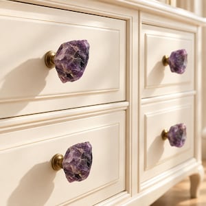 May include: A set of four drawer knobs featuring raw, deep purple amethyst crystal stones attached to circular brass-toned metal bases. The knobs are mounted onto a cream-coloured wooden dresser with rectangular recessed panels on each drawer front.