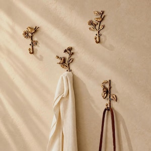 May include: Four wall-mounted hooks made of antique gold-toned metal, each featuring a sculpted bird perched on a branch with leaves and flowers. One hook holds a white fabric cloth, and another hook holds a brown leather strap.