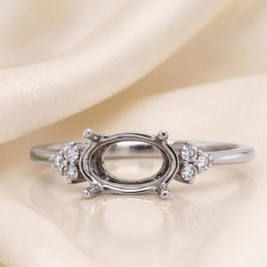 Peut inclure: Une bague en métal argenté dotée d'un sertissage ovale central vide conçu pour une pierre précieuse. L'anneau est orné de chaque côté par un groupe de trois petites pierres rondes, transparentes et étincelantes disposées en triangle.