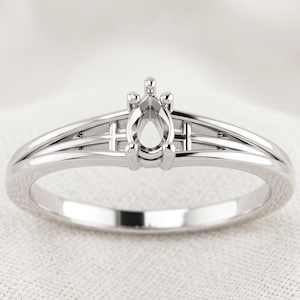 Peut inclure: Une monture de bague en métal argenté dotée d'un serti à griffes en forme de poire au centre. L'anneau se compose de deux rails métalliques parallèles qui convergent vers la tête, créant une structure architecturale ouverte pour une pierre.
