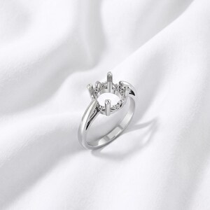 Könnte beinhalten: Eine Ringfassung aus silberfarbenem Metall mit einem kreisförmigen Kopf und vier Krappen, die zur Aufnahme eines Edelsteins ausgelegt ist. Die Ringschiene ist glatt und poliert und mit einer filigranen, durchbrochenen Galerie verbunden.