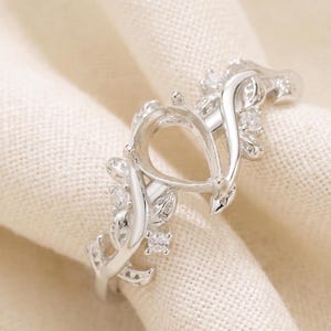 May include: A silver-coloured ring featuring a central pear-shaped prong setting designed to hold a gemstone. The band is crafted with a vine-like motif, incorporating small, clear, round-cut stones embedded within the metal leaves along the shank.