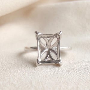 Könnte beinhalten: Ein Ring aus silberfarbenem Metall mit einer rechteckigen Fassung, vier Krappen und einem filigranen, blumenartigen durchbrochenen Design unter der Fassung, das dazu dient, einen einzelnen rechteckigen Edelstein sicher und erhöht zu halten.