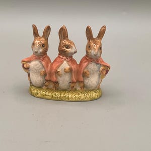 May include: A ceramic figurine featuring three brown rabbits standing in a row on a textured green base. Each rabbit has white fur on its chest and wears a pink cape tied at the neck. The rabbits are depicted with pointed ears and brown details.