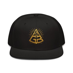 May include: A black snapback cap featuring a gold-coloured graphic on the front. The design depicts a pyramid with an eye at the top and the letters GB integrated into the base, surrounded by radiating lines, creating a distinct metallic appearance.