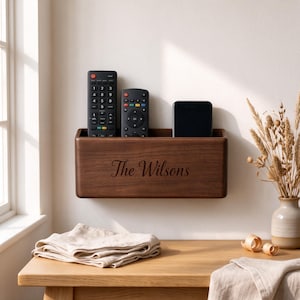 Peut inclure: Une boîte de rangement murale rectangulaire en bois foncé avec le texte gravé "The Wilsons" sur le devant. La boîte contient deux télécommandes noires et un smartphone noir, offrant un organisateur fonctionnel pour les petits objets.