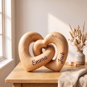 Op de afbeelding: Een lichtgekleurd houten sculptuur bestaande uit twee in elkaar grijpende hartvormen. Het hout heeft een zichtbaar nerfpatroon. De namen Emma en Jack zijn in een donker scriptlettertype gegraveerd, samen met de datum 14.02.2025.
