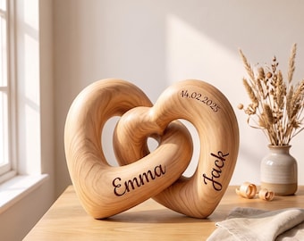 May include: A light-coloured wooden sculpture consisting of two interlocking heart shapes. The wood features a visible grain pattern. The names Emma and Jack are engraved in a dark script font, along with the date 14.02.2025 on the right heart.